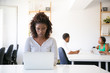 © Mangostar - Serious excited African American employee working on computer in office. Her colleagues talking at meeting table in background. Workgroup in office concept