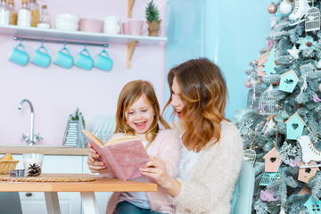 Smiling woman and little girl child are sitting at table in light christmas kitchen. Happy mother and daughter are reading book with fairy tales. Family spend time together on new year eve.