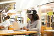 © Mangostar - Focused black female student describing VR experience in public library. Woman with taken off virtual reality glasses, sitting at desks and using laptop. VR experience concept