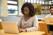 © Mangostar - Focused student working in library computer class. Young black woman sitting at desk and using laptop in classroom with bookshelves. Education concept
