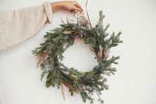 Hipster Girl In White Sweater Holding Rural Christmas Wreath With Fir Branches, Berries, Pine Cones And Herbs  On White Wall In Room. Christmas Rustic Wreath. Season's Greetings