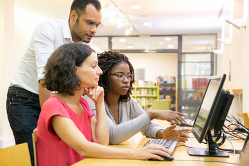 Wall Mural - tutor helping students in computer class. man and women sitting and standing at desk, using desktop,