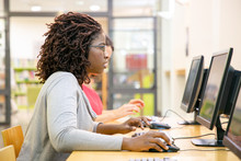 Focused Female Student Excited With Online Test In Computer Class. Black Woman In Casual Sitting At Table, Using Desktop, Typing, Looking At Monitor With Open Mouth. Excites Student Concept
