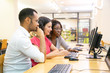 © Mangostar - Diverse team of adult students working together in computer class. Man and women in casual sitting at table, using desktop, watching content on monitor and talking. Collaboration concept