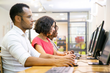 Wall Mural - Mix raced adult students training in computer class. Row of man and women in casual sitting at table, using desktops, typing, looking at monitor. Staff training concept