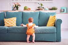 Ten Month Old, Infant Baby Girl In Yellow Bodysuit Is Standing, Leaning On Sofa At Home