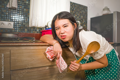 young tired and stressed Asian Chinese woman in cook apron working leaning on kitchen sink lazy in moody and upset face housekeeping at home and crying