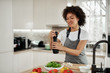 © nenadaksic - Attractive mixed race woman in gray apron addinf pepper in bowl with rocket and mushrooms. On kitchen counter are different vegetablet.
