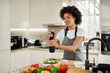 © nenadaksic - Attractive mixed race woman in gray apron addinf pepper in bowl with rocket and mushrooms. On kitchen counter are different vegetablet.