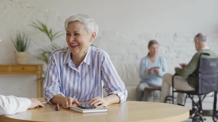 Wall Mural - Tracking shot of elderly couple sitting at table and having fun talking joyfully, senior woman and disabled senior man in wheelchair playing cards in background in assisted living home