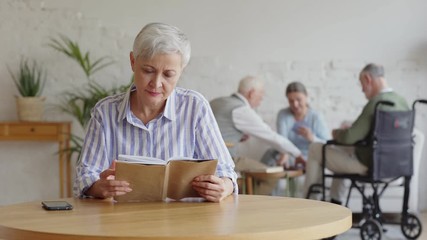 Wall Mural - Rack focus of three elderly people, two men including disabled one and woman, playing cards in nursing home. Old woman with short grey hair reading book and looking at camera sitting at table