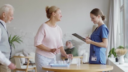 Wall Mural - Group of senior patients taking vitamin pills in nursing home and thanking young female nurse in blue uniform making marks in list on clipboard. Elderly people gathering and talking in background