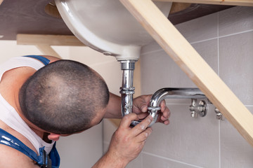 Wall Mural - Closeup male plumber worker in blue denim uniform, overalls, fixing sink in bathroom with tile wall. Professional plumbing repair service, installation water pipes, man mounted sewer drain