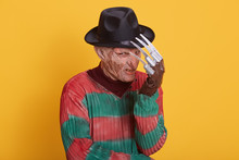 Studio Portrait Of Man In Monster Makeup, Having Long Hose And Ugly Face, Freak Hiding Behind His Hand, Has Knives Instead Of Fingers, Wearing Striped Sweater With Holes And Hat, Yellow Background.