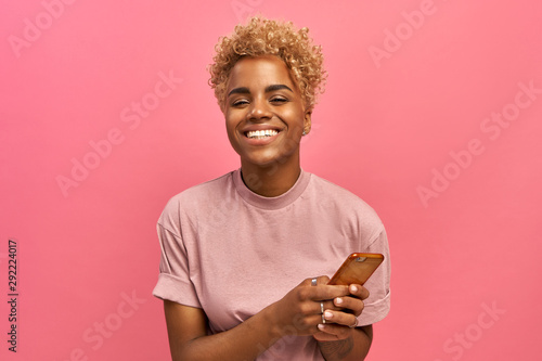 Half length shot of positive attractive female model with Afro haircut, feels good, uses smartphone device for entertainment and online chatting, surfers social network profile, uses free internet