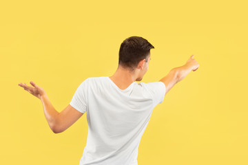 Caucasian young man's half-length portrait on yellow studio background. Beautiful male model in shirt. Concept of human emotions, facial expression, sales, ad. Showing and pointing something.