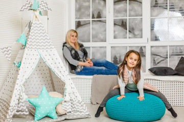 Wall Mural - A young mother in a gray sweater and daughter are sitting in the children's room in winter. the Christmas and New Year's interior. Holiday. Family concept.