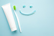 Toothbrush with green bristles, white tube of toothpaste on pastel blue background. Happy, smiley face created from paste. Healthy teeth concept. Empty place for text.