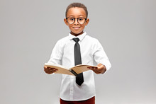 Studio Image Of Funny Smart Akward African Schoolboy In Formal Clothes And Eyeglasses Eager To Learn, Holding Open Book In His Hands And Smiling. Enthusiastic Dark Skinned A-student Learning At School
