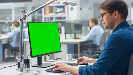 Leinwandbilder - Over the Shoulder Shot of Engineer Working with Green Mock-up Screen Desktop Computer. In the Background Engineering Facility with Specialists Working on Blueprints and Drawings with Industrial Design