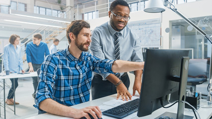 Leinwandbilder - In the Industrial Engineering Facility: Portrait of the Handsome Male Engineer Working on Desktop Computer, Chief Engineer Stands Beside and Explains Particularities of the Task and Project Details. 