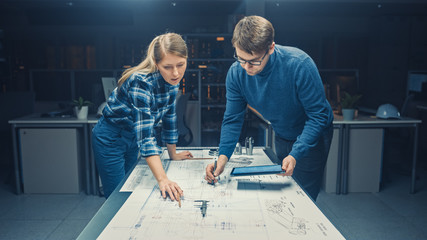 In the Dark Industrial Design Engineering Facility Male and Female Engineers Talk and Work on a Blueprints Using Conference Table. On the Desktop Drawings, Drafts and Electric Engine Components, Parts