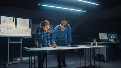 Wall Mural - In the Dark Industrial Design Engineering Facility Male and Female Engineers Talk and Work on a Blueprints Using Digital Tablet and Conference Table. On the Desktop Drawings and Engine Components