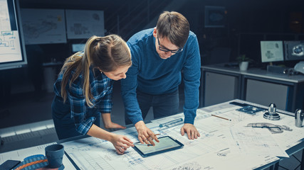 Wall Mural - In the Dark Industrial Design Engineering Facility Male and Female Engineers Talk and Work on a Blueprints Using Digital Tablet and Conference Table. On the Desktop Drawings and Engine Components