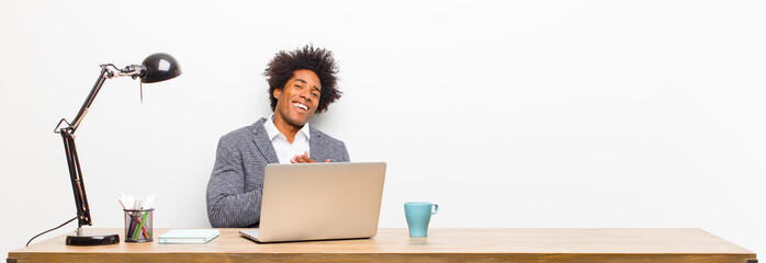 Wall Mural - young black businessman feeling romantic, happy and in love, smiling cheerfully and holding hands close to heart on a desk