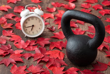 Black Iron Kettlebell On A Rustic Wood Floor, With White Analog Alarm Clock And Red Fall Maple Leaves
