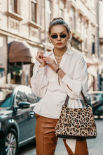 Outdoor Autumn Fashion Portrait Of Elegant, Luxury Woman Wearing Sunglasses, Trendy White Shirt, Wrist Watch, Leather Trousers, Holding Animal, Leopard Print Bag, Posing In Street Of European City