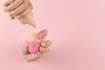 Sticker - Girl hand holds a beauty blender in foundation, concealer from a hole in a pink background. Makeup artist concept, copy space.