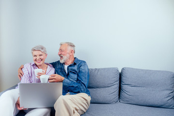 elderly couple in love sitting gray couch in living room look at screen laptop and drinking cup of tea or coffee copyspase