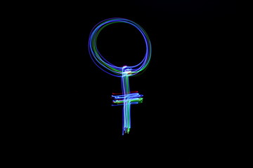 Long exposure photograph of a female symbol in neon colour in an abstract swirl, parallel lines pattern against a black background. Light painting photography.