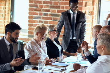 Wall Mural - Group of businessmen wear suits sit at table in office and discuss new project, smile. caucasian blond woman and african man stand, behind open window. Everyone move, work.