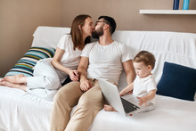 Loving Couple Kissing While Their Baby Watchcing Cartoon On The Laptop, Love, Warm, Tender Feeling And Emotion