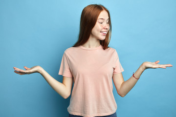 positive happy woman raises both palms, pretends holding two items, being in good mood, wears pink T-shirt, isolated over blue background. Satisfied student holds copy space. close up portrait