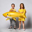 © FAB.1 - Positive little children, dressed in festive clothes, wears a balloon in the shape of a yellow shark fish, celebrates a holiday, has a wide smile, stands on a light background, being in a good mood.