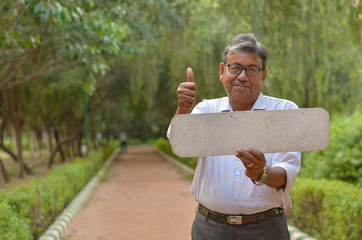 Senior Bengali Indian man holding a blank placard showing thumbs up which can be used for quotes in a park in New Delhi, India. Concept motivational