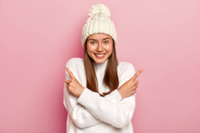 Positive Young Girl With Straight Hair, Crosses Arms And Points In Both Sides, Wears Fashionable White Hat And Jumper, Chooses Between Two Items, Smiles Pleasantly, Isolated Over Pink Background.