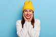 © WHstudio Leushin N - Pretty happy European woman wears red lipstick, keeps hands on cheeks, wears yellow hat and white cashemere sweater, poses against blue background. Cheerful lovely feminine girl feels amused
