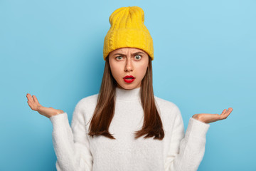 Wall Mural - Portrait of beautiful young Caucasian girl being questioned, spreads palms sideways, feels unawareness and doubt, wears red lipstick, wears yellow stylish hat, white jumper, isolated on blue