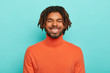 Carefree smiling dark skinned millennial guy has happy facial expression, laughs at something positive, shows white teeth, wears orange poloneck, enjoys spare time, has dreadlocks, poses indoor
