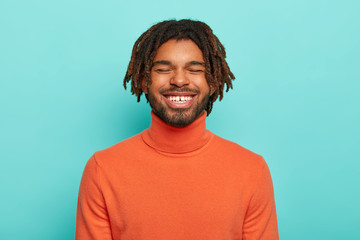 Wall Mural - Carefree smiling dark skinned millennial guy has happy facial expression, laughs at something positive, shows white teeth, wears orange poloneck, enjoys spare time, has dreadlocks, poses indoor