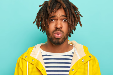 Wall Mural - Headshot of displeased dark skinned male looks surprisingly at camera, wears raincoat as walks in rainy weather, has short dreadlocks, puzzled by shocking relevation, isolated over blue background