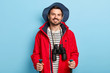 Positive male traveler uses trekking sticks for walking in forest, spends vacation actively, smiles positively, dressed in stylish headgear and red jacket, has binoculars on neck. Travel lifestyle