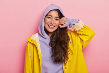 Wall Mural - Positive brunette Korean girl in casual hoody and raincoat, smiles pleasantly, being in good mood after outdoor walk with boyfriend, stands against pink background. Cold rainy autumn weather