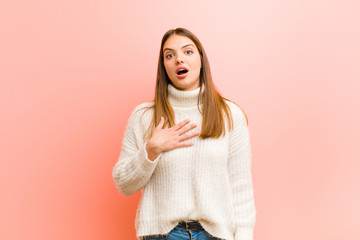 young pretty woman feeling shocked, astonished and surprised, with hand on chest and open mouth, saying who, me? against pink background