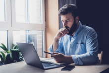 Professional Sitting In Office In Front Of Laptop. Developer Thinking On Solutions For Work. Home-based Student Getting Distant Education. Young Serious Bearded Man In Blue Shirt Working On Desktop.