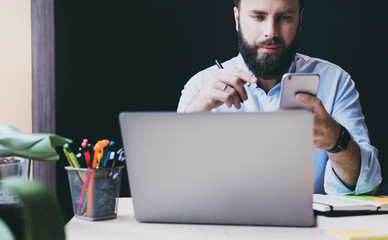 Professional sitting at office table in front of laptop and using smartphone. Male person sending text messages and checking email on mobile phone. Digital online solutions and apps for office work.